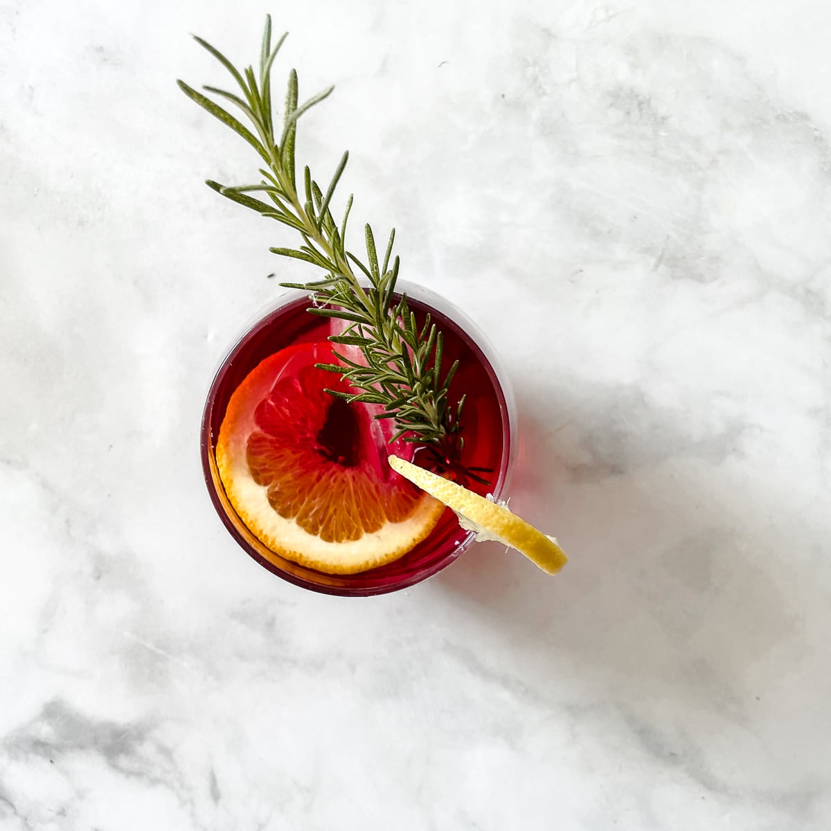 A cranberry margarita in a glass with an orange slice and lemon and garnished with rosemary.