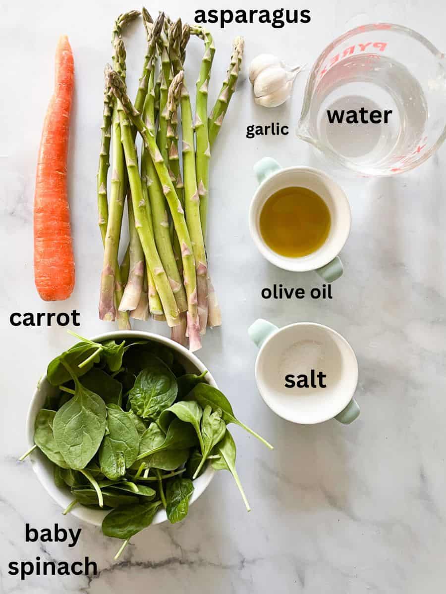 Creamy vegan asparagus soup that's full of fresh vegetable flavors and vibrantly green from plenty of asparagus. Ingredients for vegan asparagus soup are shown labelled and portioned out: asparagus, carrot, salt, olive oil, water, spinach, and garlic.