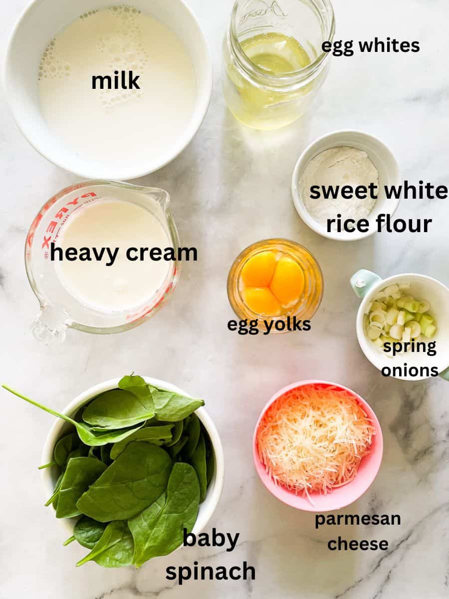 The fluffiest gluten free souffle with spinach and parmesan cheese is easily made gluten-free by using sweet white rice flour. The ingredients for gluten free souffle are shown labeled and portioned out: egg whites, egg yolks, sweet white rice flour, milk, cream, spinach, parmesan cheese, spring onions.