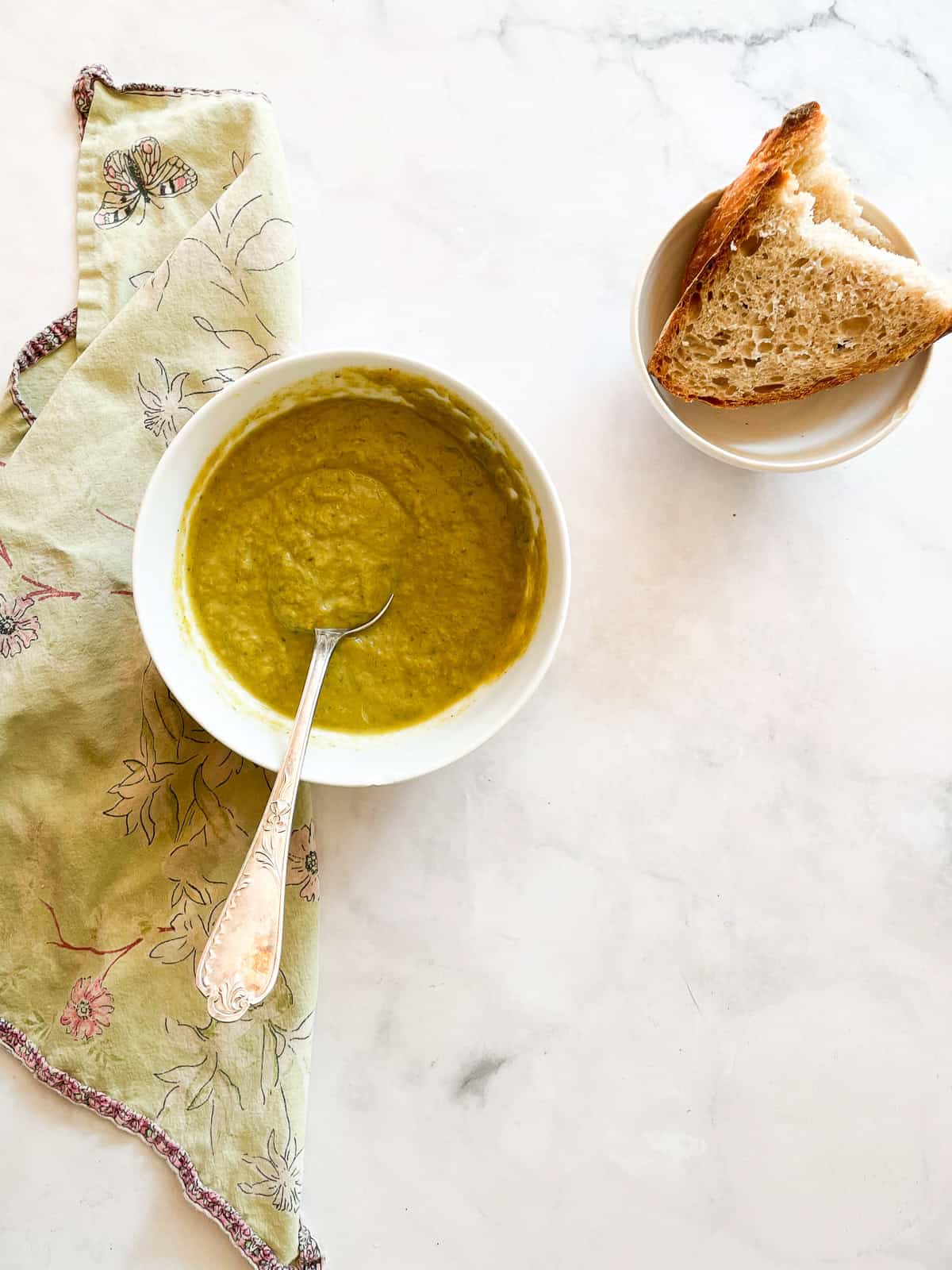Creamy, cozy asparagus potato soup is crammed with vibrant vegetables like carrots, spinach, and potatoes. Pretty and satisfying! A bowl of asparagus potato soup with a spoon in it next to a bowl of sliced bread.