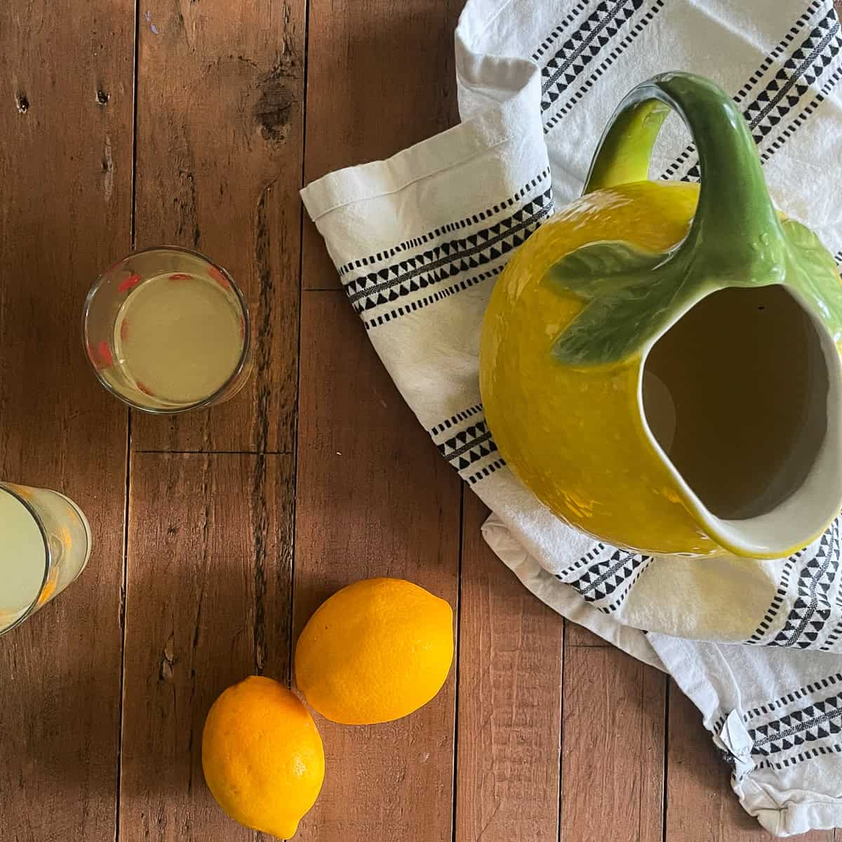 A pitcher of homemade lemonade with lemons and glasses of lemonade.