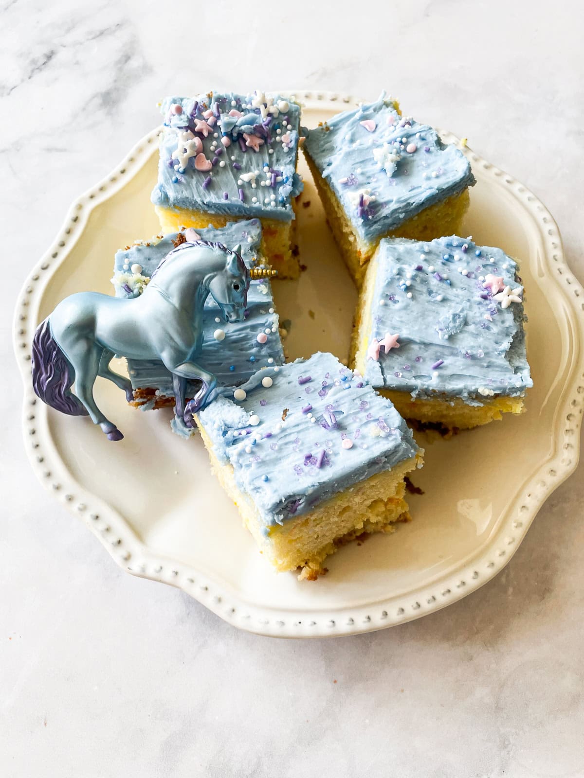 These adorable unicorn bars are perfect for a kid's birthday party! A buttermilk funfetti sheet cake is topped with blue icing and plenty of sprinkles. A unicorn stands on a plate of unicorn bars.