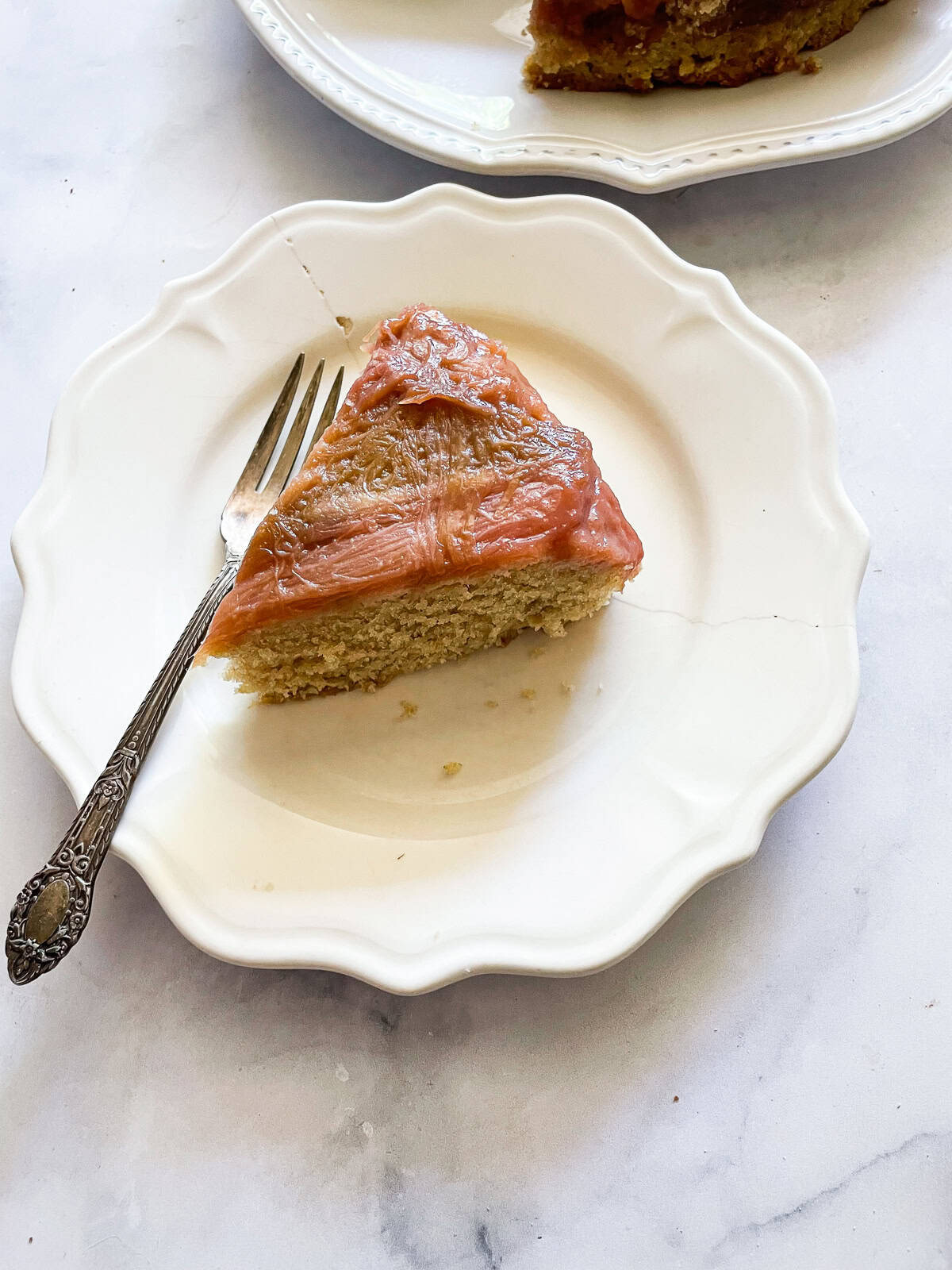 This gorgeous upside-down rhubarb cake drapes buttery rhubarb over a vanilla-scented cake for a beautiful, summery treat. A slice of upside-down rhubarb cake on a plate next to a cake.