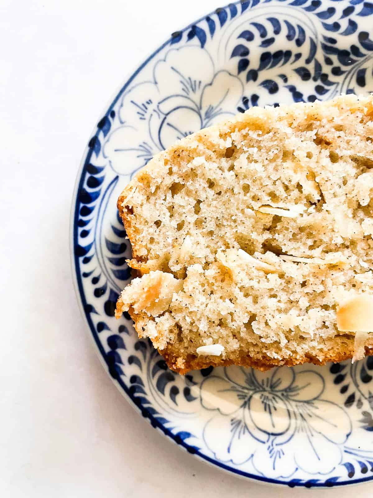 A cinnamon-kissed coconut loaf cake packed with bits of flaked coconut. Inspired by the classic Bill Granger recipe from "Australian Food." Half of a slice of coconut loaf cake on a plate.
