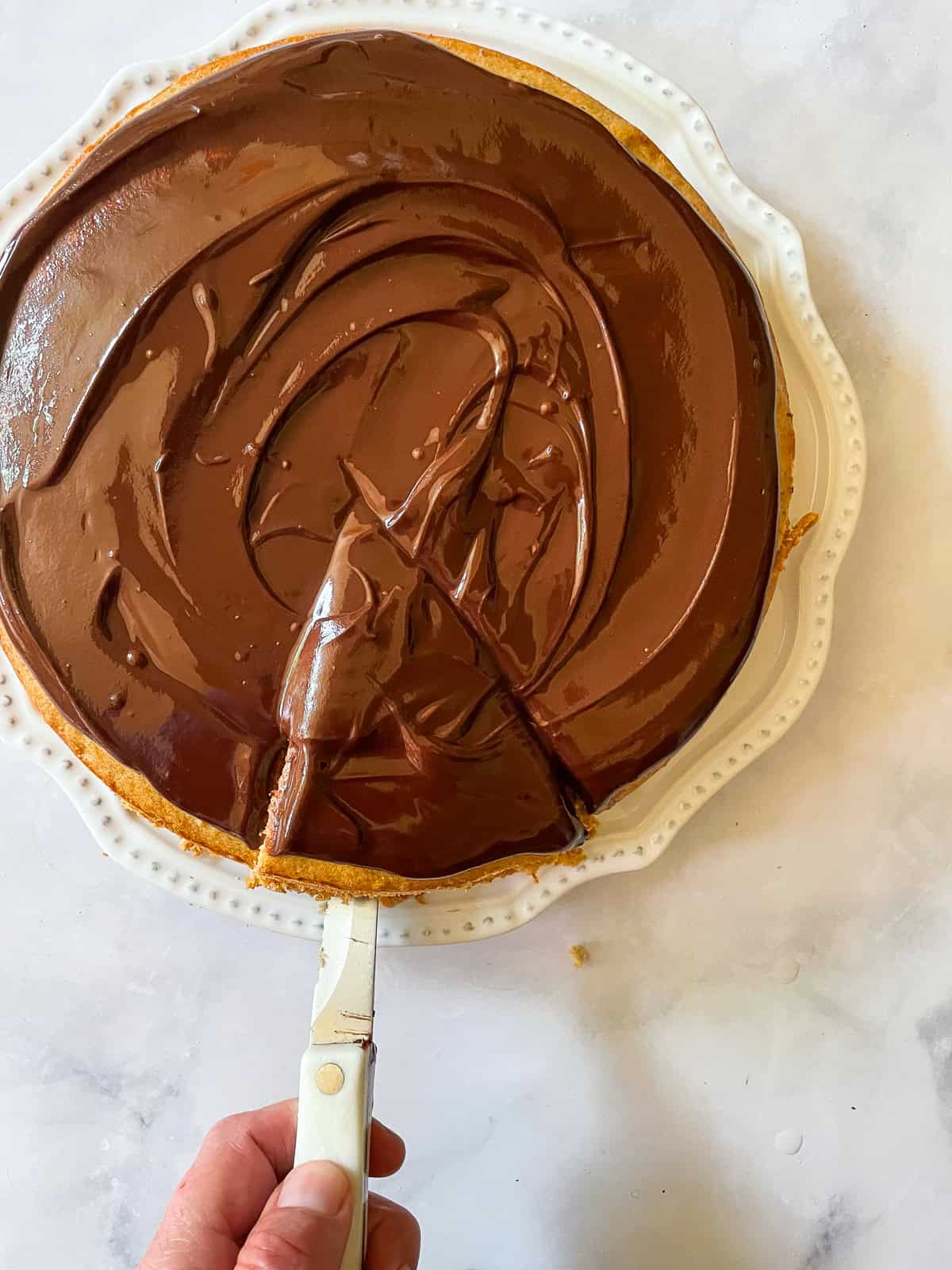 A hand uses a knife to cut a piece of banana cake with chocolate frosting.