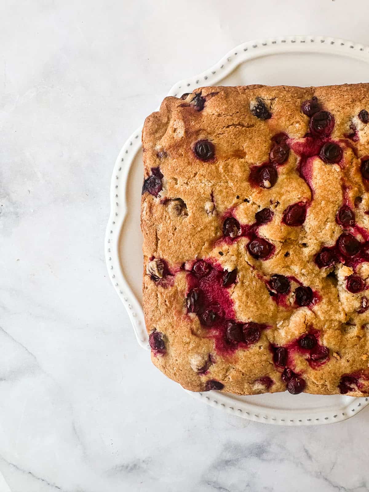 Studded with juicy berries, this sweet and soft cranberry cake is a lovely holiday bake. Great with brunch, or as dessert with ice cream. Half of a cranberry cake is seen on a serving plate.