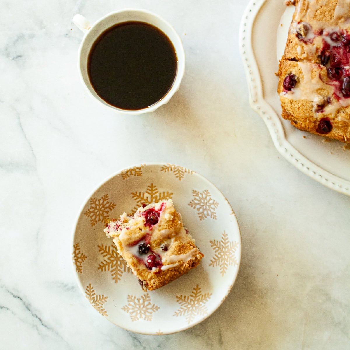 Studded with juicy berries, this sweet and soft cranberry cake is a lovely holiday bake. Great with brunch, or as dessert with ice cream. A piece of cranberry cake on a plate with a cup of coffee.