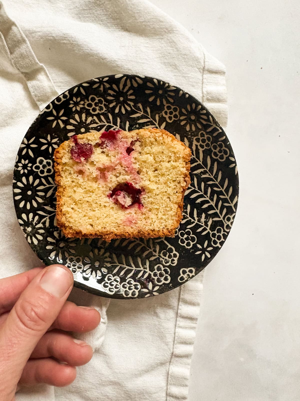 This gorgeous lemon cranberry bread features a lemon-scented loaf swirled with juicy cranberries. Perfect for holiday baking. A hand reaches for a plate with a slice of lemon cranberry bread.