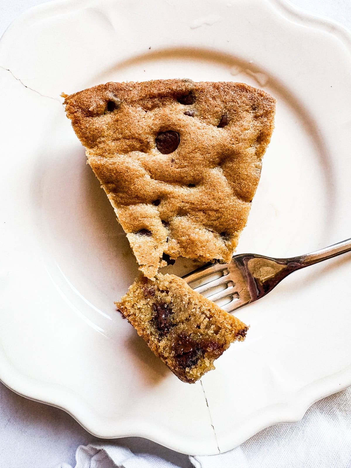 This buttery chocolate chip cookie cake is made with whole wheat flour and brown sugar for a touch of caramel sweetness. A fork cuts into a slice of chocolate chip cookie cake on a plate.