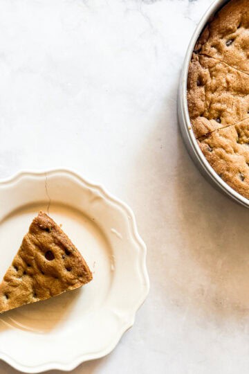 A slice of chocolate chip cookie cake on a plate with the sliced cake to the right of it.