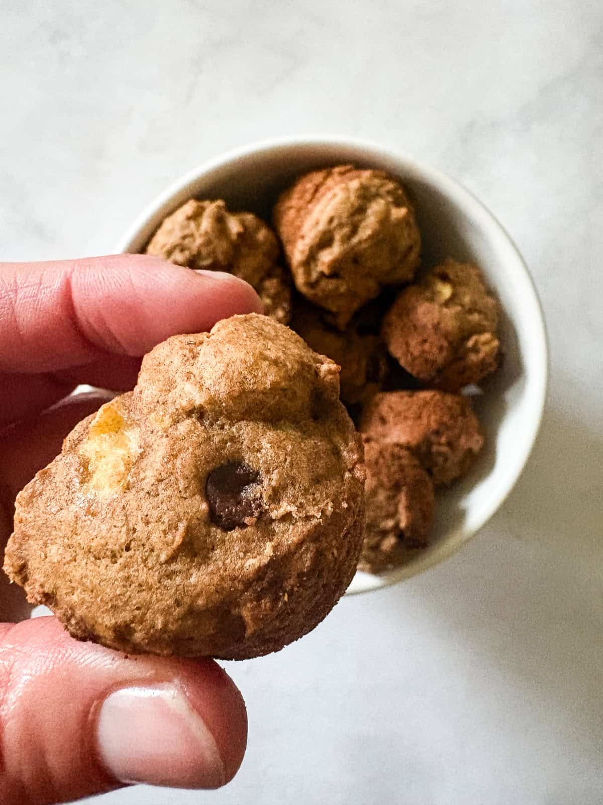 These perfect bite-sized mini banana muffins are made with whole-grain flour, maple syrup, and heart-healthy olive oil. A hand holds a mini banana muffin over a bowl of muffins.