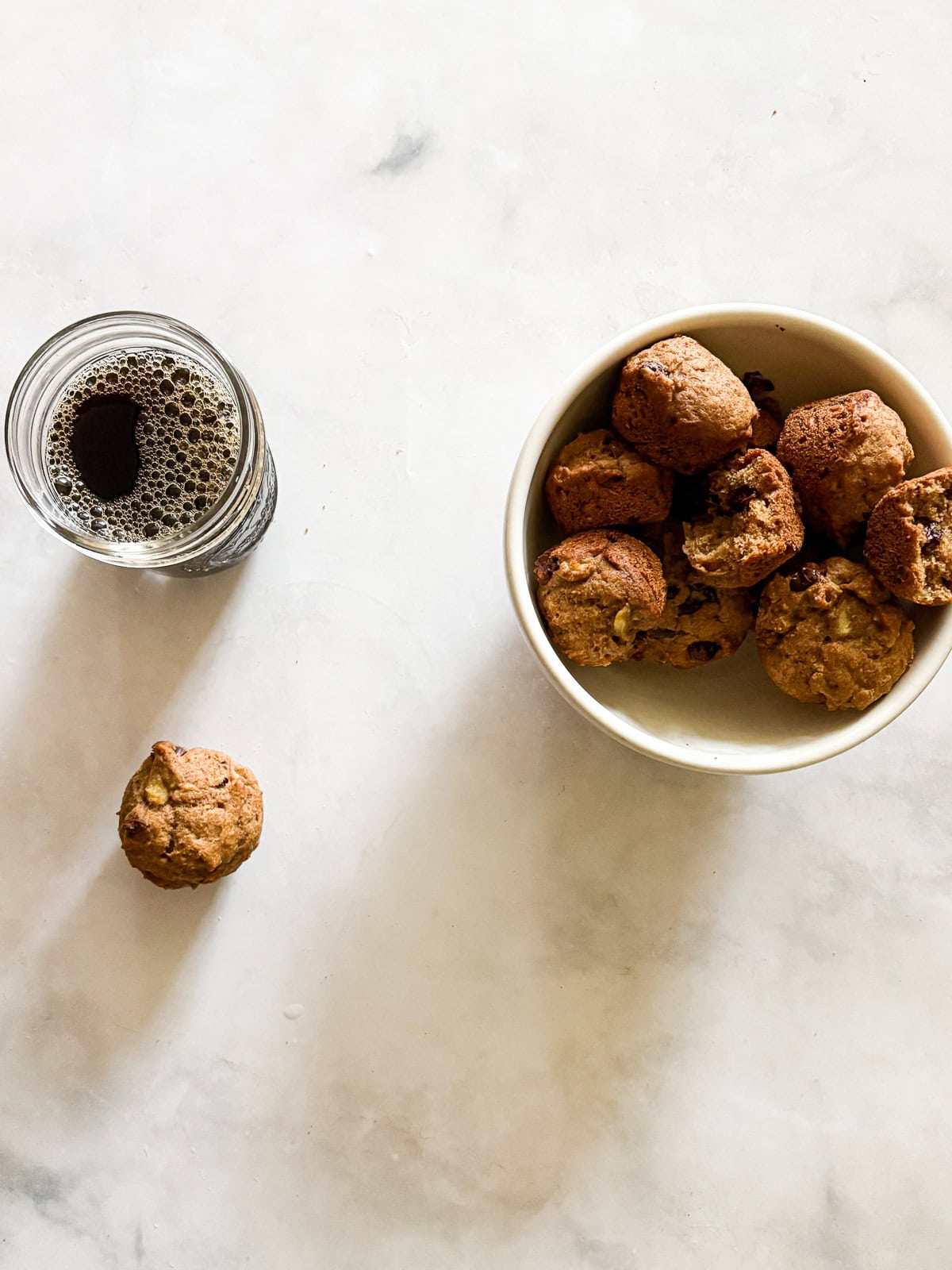 These perfect bite-sized mini banana muffins are made with whole-grain flour, maple syrup, and heart-healthy olive oil. A bowl of mini banana muffins and a jar of coffee.