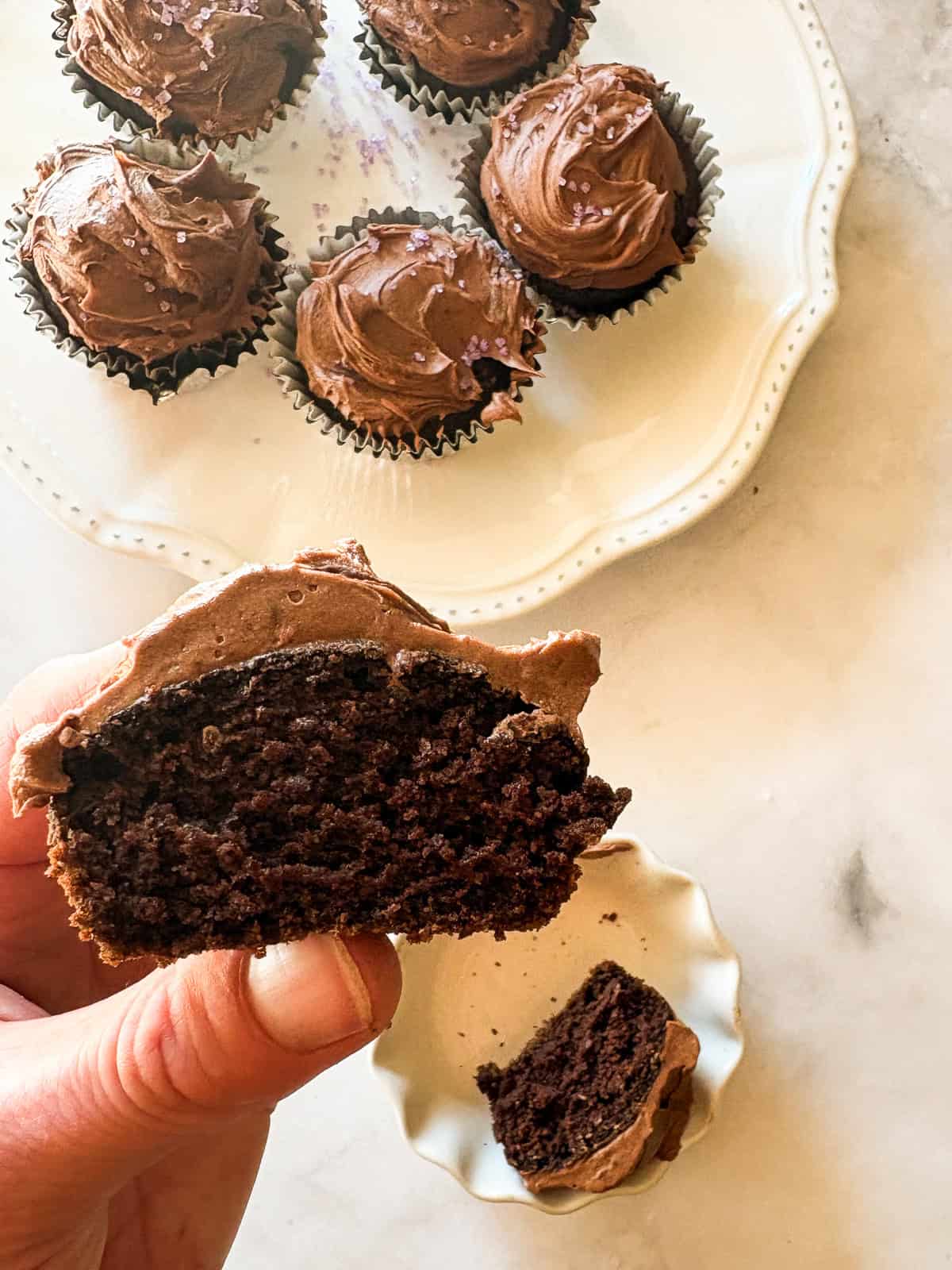 Tender, chocolatey vegan chocolate cupcakes frosted with a sumptuous vegan chocolate buttercream are out-of-this-world good! A hand holds up a vegan chocolate cupcake that has been cut in half.